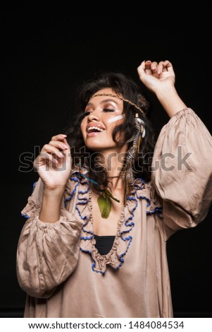 Portrait of a beautiful cheerful young brunette woman wearing tribal clothing and accessories standing isolated over black background, dancing
