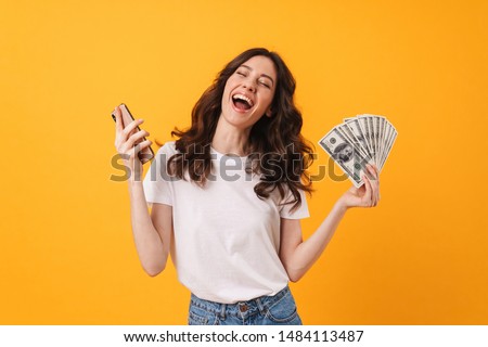 Photo of pleased happy emotional screaming young woman posing isolated over yellow wall background using mobile phone holding money.