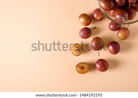 Scattered sliced half ripe sweet plum fruits with water drops near to plums in glass bowl on cream colored background, bright, top view, flat lay