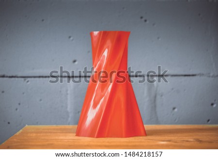 Bright red object vase printed by 3d printer on blue brick wall background. Automatic three dimensional 3d printer performs plastic red colors modeling in laboratory. Modern 3D printing technology.