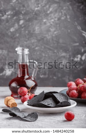 Black potato chips with charcoal, balsamic vinegar in glass, red grapes on a blue ceramic plate on a gray concrete background. side view, close up, selective focus, copy space.