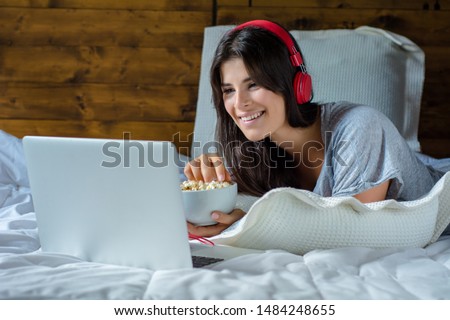 Young woman using laptop, eating popcorn and watching movies in bed at home. Royalty-Free Stock Photo #1484248655