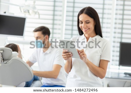 smiling female dentist using digital tablet while her collegue working with small parient on the background