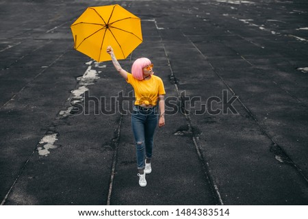 Young Asian millennial hipster in stylish outfit and pink wig carrying bright yellow umbrella and looking away while walking on weathered ground on city street Royalty-Free Stock Photo #1484383514
