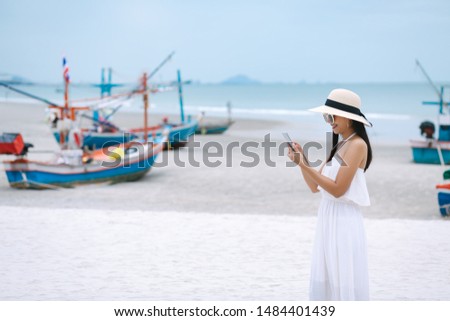 Young travel asian woman with white dress using mobile phone on beach against sea fishing boat background in summer, Thailand