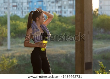 Slim athletic girl takes a break between classes and drinks water from a bottle, urban background. Beautiful sky at sunset.