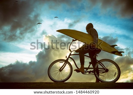 Silhouette of surfer carrying his surfboard on a beach cruiser bike in front of bright skyscape