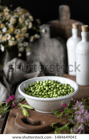 Young green peas in white bowl on wooden background. Pea flowers and daisy flowers on the table. Dark photo