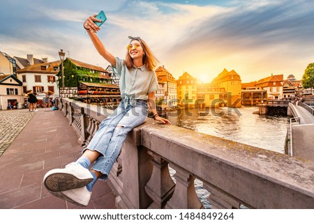 Happy and cheerful Asian girl traveler takes a selfie on the Saint Martin bridge in the Petit France area in Strasbourg. Colorful and dramatic sunset at the background