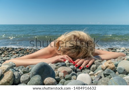 Blonde girl lying on her stomach on a pebble beach near the sea on a Sunny day; in the background the sea, the sky.
