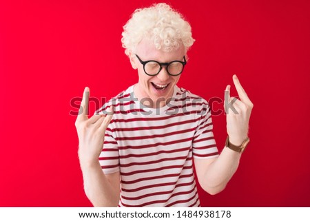 Young albino blond man wearing striped t-shirt and glasses over isolated red background shouting with crazy expression doing rock symbol with hands up. Music star. Heavy concept.