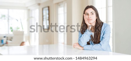 Wide angle picture of beautiful young woman sitting on white table at home thinking looking tired and bored with depression problems with crossed arms.