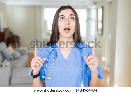Beautiful young nurse woman wearing uniform and stethoscope at the clinic amazed and surprised looking up and pointing with fingers and raised arms.