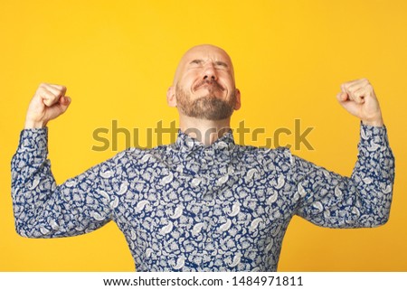 Fabulous at any age. Portrait of 40-year-old man feeling successful over light yellow background in blue shirt. Hands up. Hipster style. Bald shaved head. Text space. Studio shot