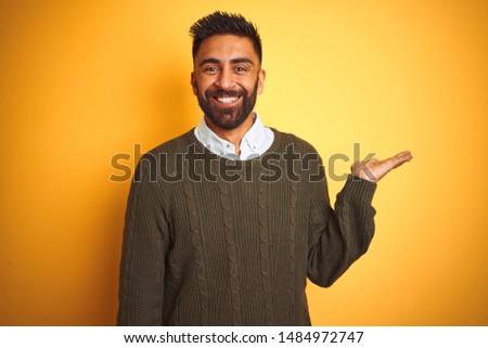 Young indian man wearing green sweater and shirt standing over isolated yellow background smiling cheerful presenting and pointing with palm of hand looking at the camera.