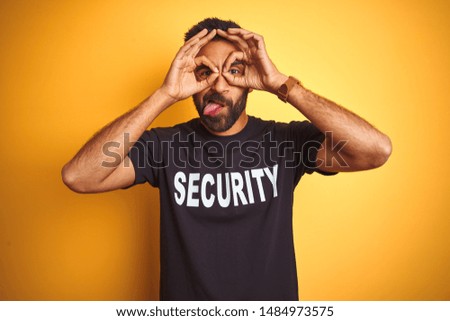 Arab indian hispanic safeguard man wearing security uniform over isolated yellow background doing ok gesture like binoculars sticking tongue out, eyes looking through fingers. Crazy expression.
