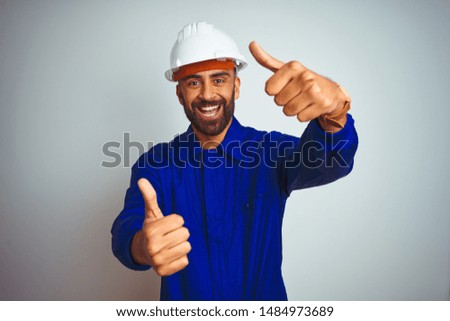 Handsome indian worker man wearing uniform and helmet over isolated white background approving doing positive gesture with hand, thumbs up smiling and happy for success. Winner gesture.