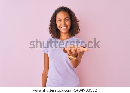 Young brazilian woman wearing t-shirt standing over isolated pink background smiling cheerful offering palm hand giving assistance and acceptance.