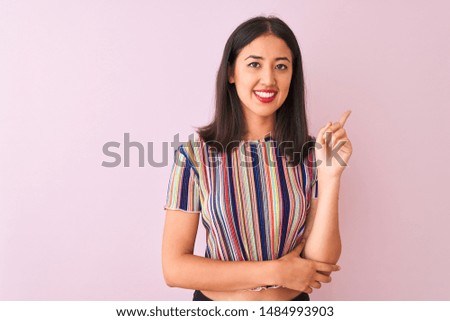 Young chinese woman wearing colorful striped t-shirt standing over isolated pink background with a big smile on face, pointing with hand and finger to the side looking at the camera.