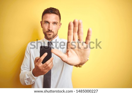 Young handsome business man using smartphone over yellow isolated background with open hand doing stop sign with serious and confident expression, defense gesture