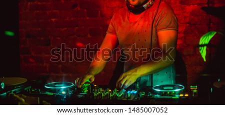 Disc jockey with cropped head in fashion grey t-shirt playing music on turned on and lightning dj deck in cafe club restaurant under color spot lights with red brick wall and a little green leaf near Royalty-Free Stock Photo #1485007052
