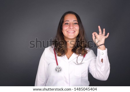 Glad attractive doctor woman shows ok sign with hand as expresses approval, has cheerful expression. Photo of beautiful female has appealing appearance, being optimistic. Standing against gray wall.