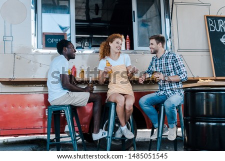cheerful multicultural men sitting and holding bottles of beer near attractive redhead woman and  food truck 