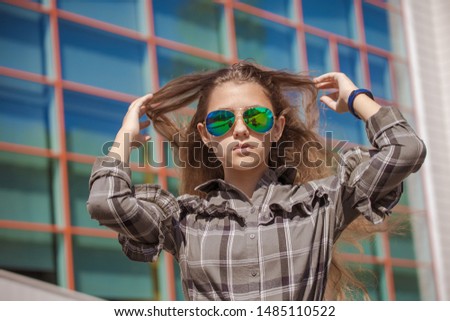 Emotional teenager girl with long hair in blue sunglasses walks around the city against the background of modern buildings