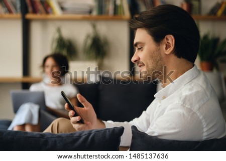 Side view of young smiling businessman sitting on sofa with colleague on background happily using cellphone in modern co-working space
