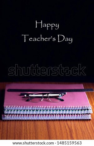 Happy Teachers' Day written on a blackboard with a stack of books or assignments, glasses, pens and ruler on a wooden table. Copy Space.