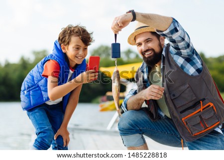 Boy taking picture. Cheerful handsome dark-haired boy taking picture of fish while fishing with dad