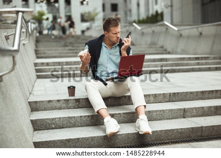young handsome office worker having unpleasant business conversation while eating his sandwich and drinking coffee for lunch during break sitting on the stairs outside