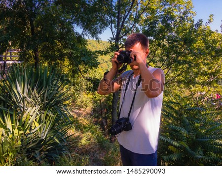 A man with a digital camera hanging on his chest photographs with a film camera against a background of green vegetation on a sunny summer day.