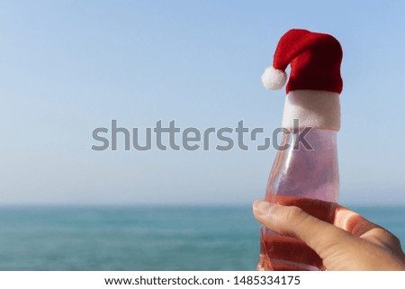 man hand with a bottle on a beach against sea with Santa Claus hat. Christmas and New Year vacation in hot country. copy space.