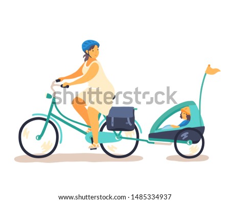 Happy woman riding a bicycle with a young girl sitting in a pull behing bike trailer. Urban family cycling and bike helmet safety topics. 
