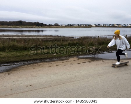 A female skateboarder in a yellow beanie and white sweatshirt pushes on her longboard as she skates along a coastal path with beach huts and a lake behind her. She cruises along an open, dirty road