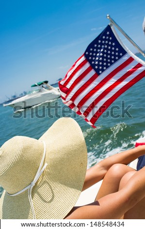 Young woman on a boat wearing a big hat with American flag waving and boat passing by.