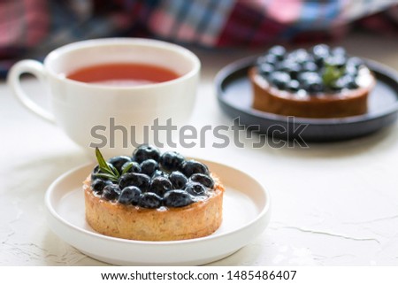 Delicious blueberry tartlets with vanilla custard cream on a black and white plates with a cup of tea on white background.  Front view. Copy-space. Selective focus