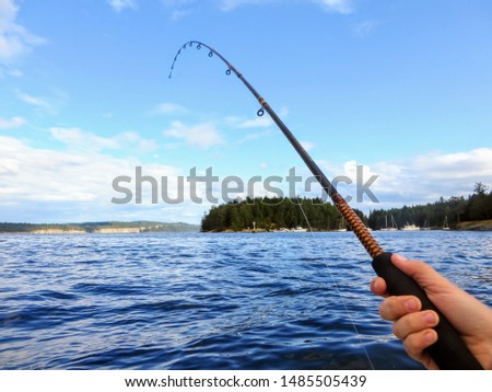 A male hand holding a fishing rod over the ocean.  The rod is pointing left and upwards towards the sky.  Room for text.