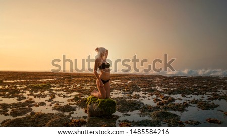 Girl in a swimsuit on a rock on her knees near the ocean at sunset