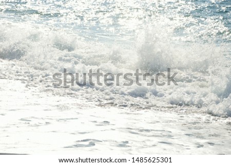 Surf on the beach with white foam on the blue water that glitters in the sunlight and upwards splashes