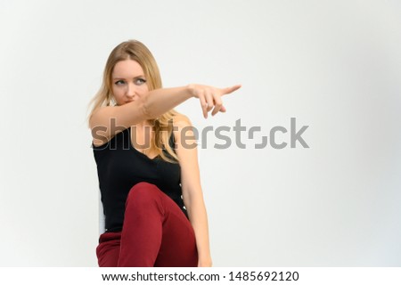 Studio waist-length photo portrait of a pretty beautiful young happy blonde woman with long hair on a white background in a black t-shirt and red trousers. Sits on a chair, smiles, shows emotions.