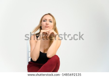 Studio waist-length photo portrait of a pretty beautiful young happy blonde woman with long hair on a white background in a black t-shirt and red trousers. Sits on a chair, smiles, shows emotions.