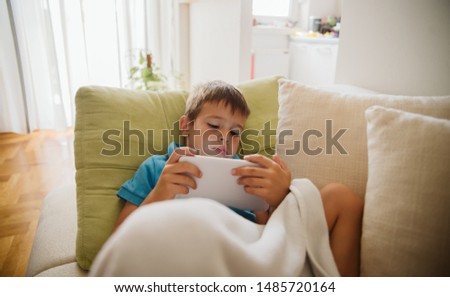 A boy in a blue T-shirt is lying on the bed and watching cartoons on a tablet