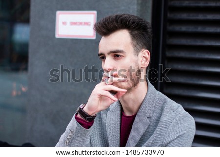 Handsome bearded young man or guy smoking a cigarette with serious thoughtful emotion smoking in prohibited place near sign no smoking. Tired and exhausted businessman in jacket or blazer takes a puff