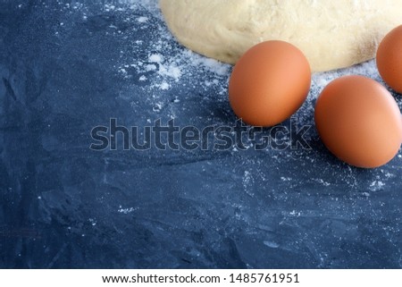 A ball of raw bread dough, sprinkled white wheat flour and three brown eggs in the corner of the picture on a blue textured concrete background with empty spaces for text and other design elements