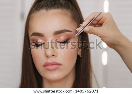Close up view of make-up artist puts false eyelashes using tweezers. Background of white folding screen
