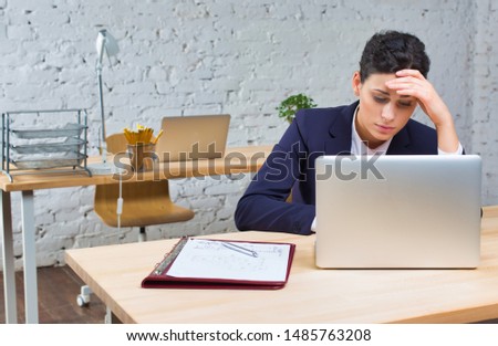Bored young businesswoman sitting with laptop at desk against brick wall in office