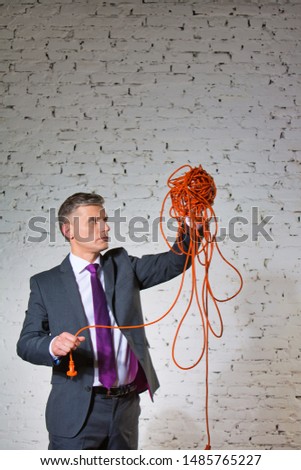 Confident mature businessman holding tangled cable against white brick wall