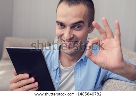 Attractive man with e-book shows okay sign, sitting on sofa at home and  looking at camera, portrait, toned